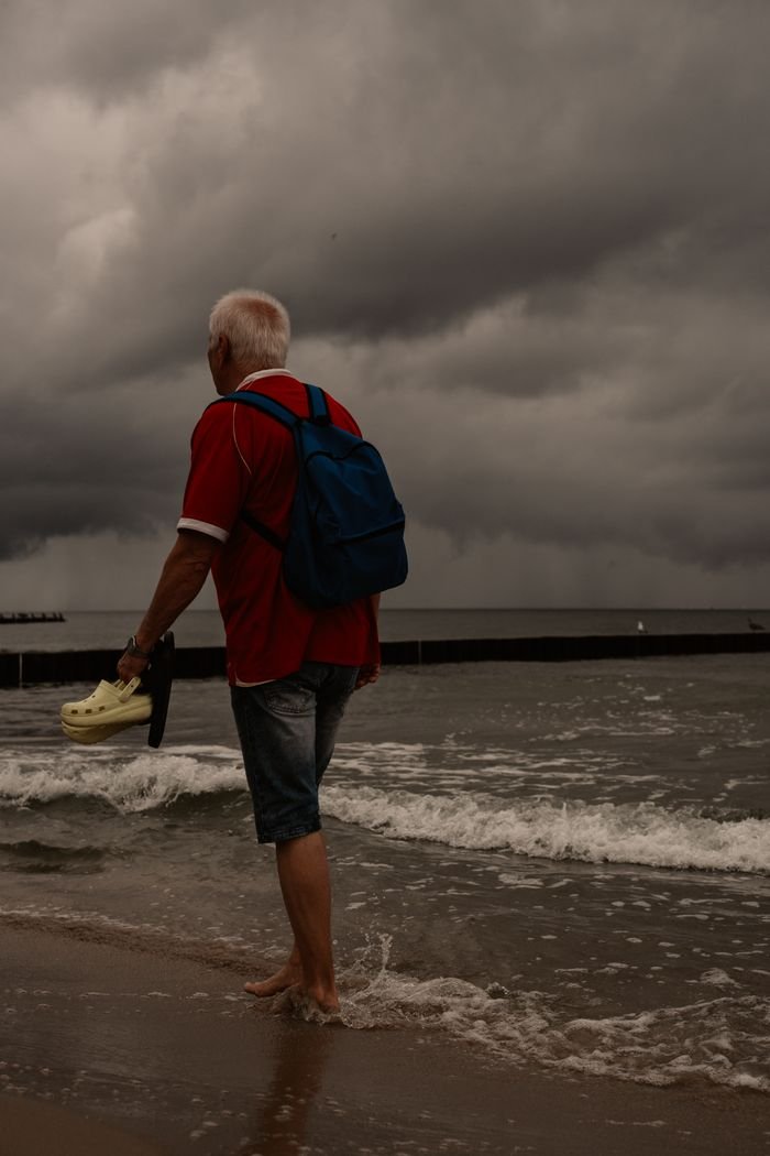 why-choose-us Senior man with a backpack strolling along a stormy beach, holding shoes as waves crash under dark clouds.