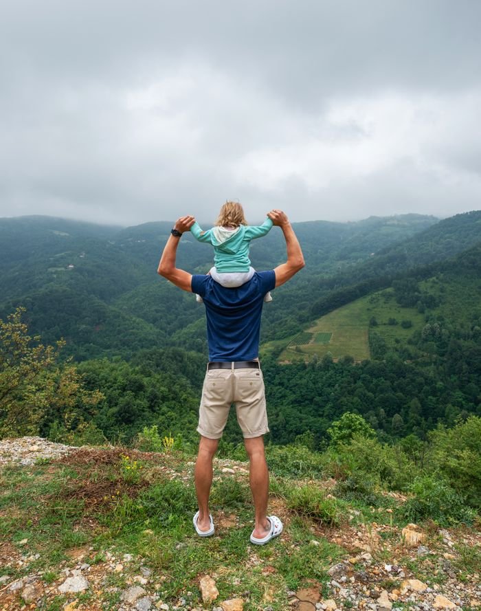 hero-img-02 Full body back view of anonymous male tourist with little unrecognizable child on shoulders standing on stony hill above grassy mountains