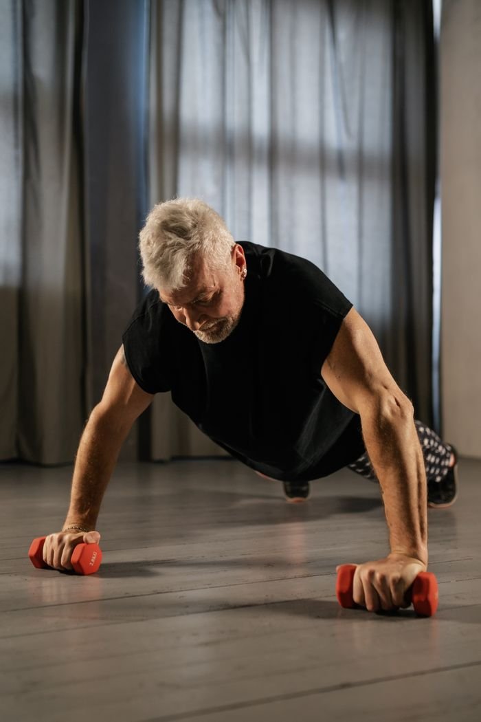 our-services-1 Elderly man performing a plank exercise indoors using red dumbbells for strength training.